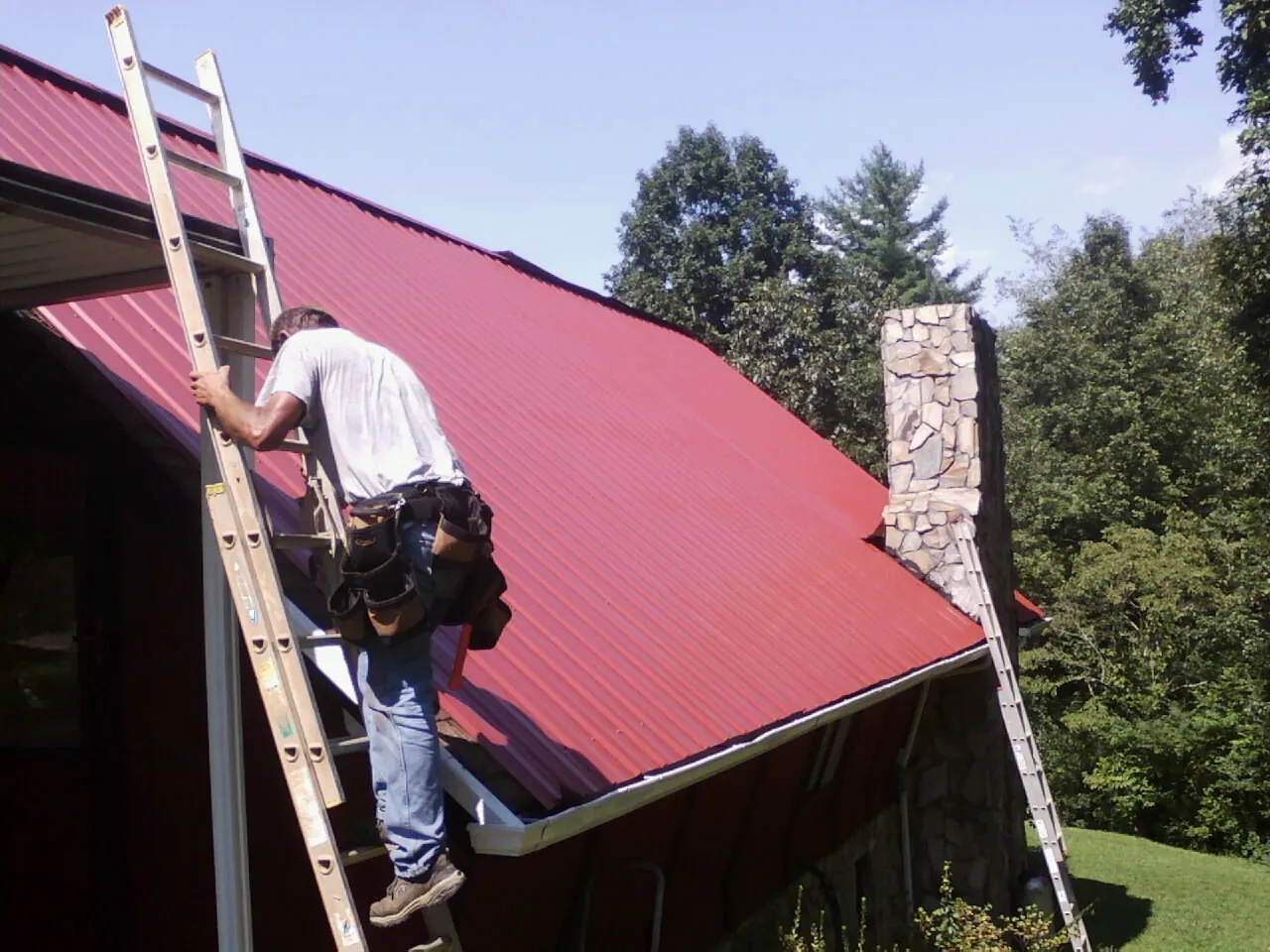 Roofer on a ladder beside a red metal roof and stone chimney — Soffit & Fascia Repair by Roofing Brothers in Abingdon